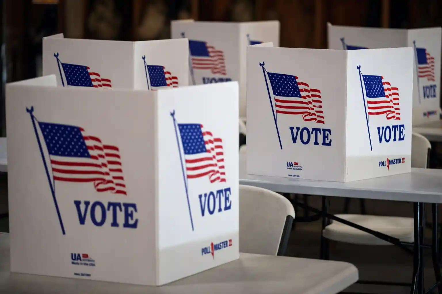 White voting booths with American flags and the word “Vote” printed on them, arranged in rows inside a polling station.