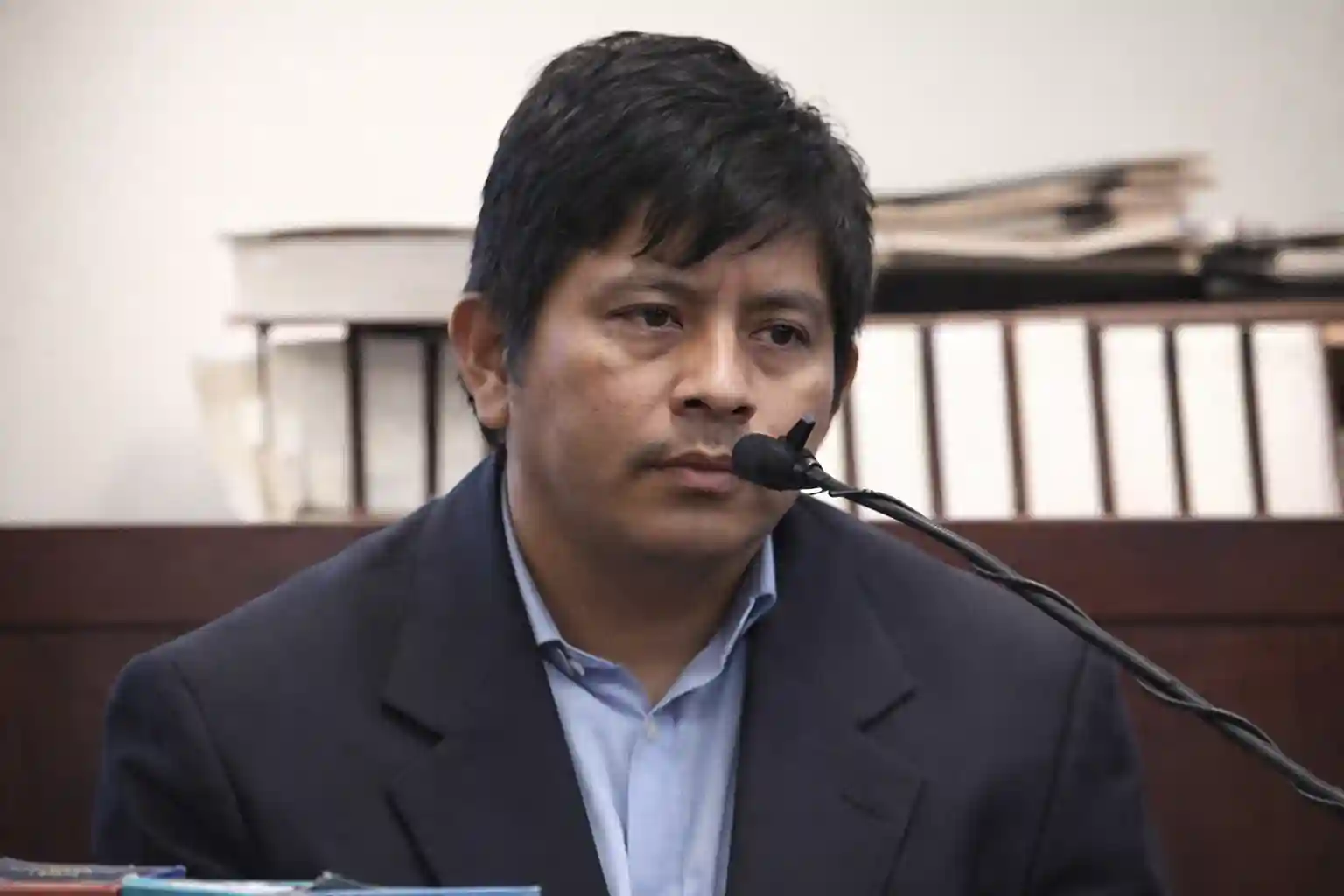 Man in a suit speaking into a microphone while seated in a courtroom.