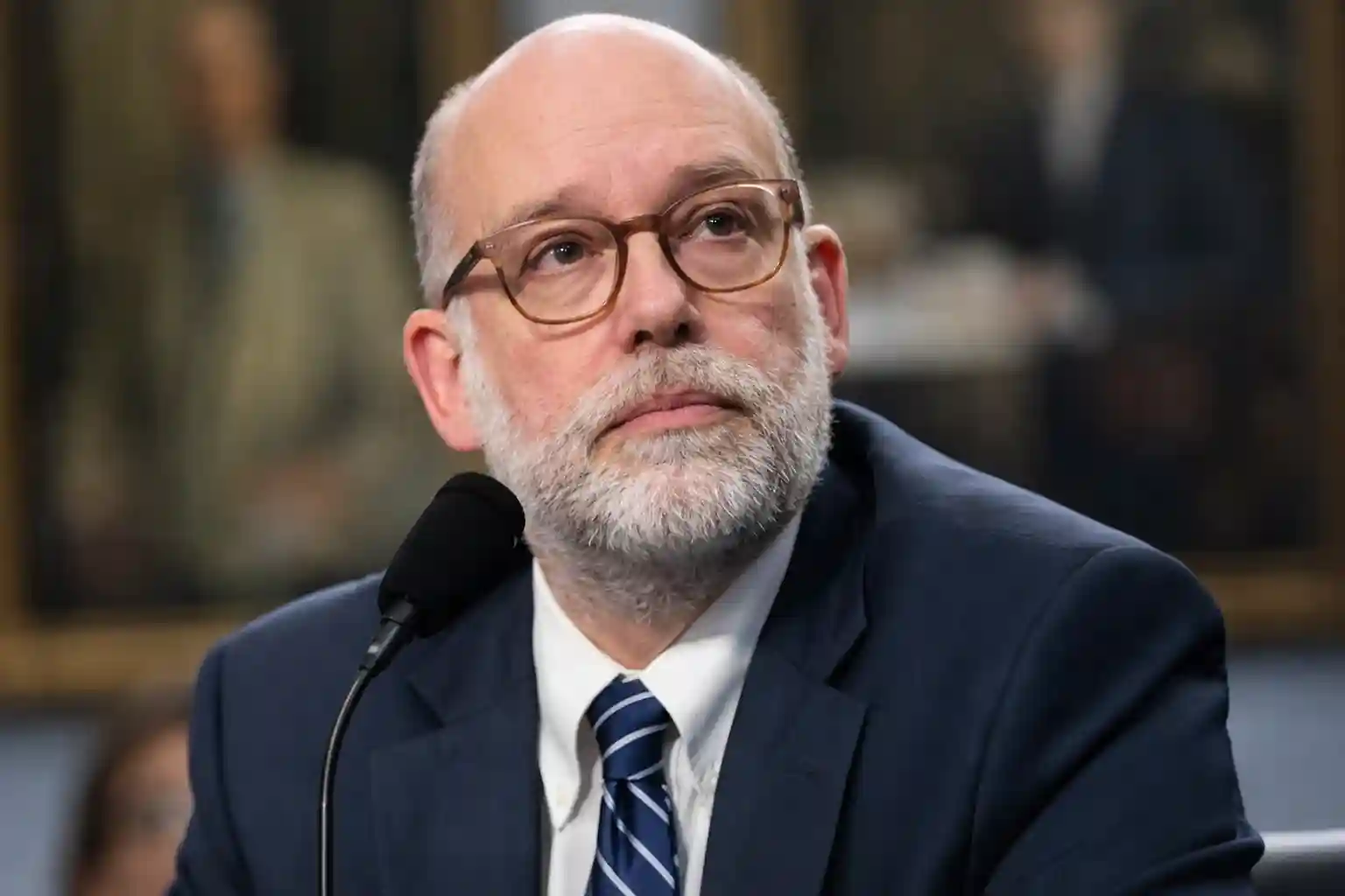 Man in a suit and glasses, speaking at a Senate hearing, looking up while listening to proceedings.