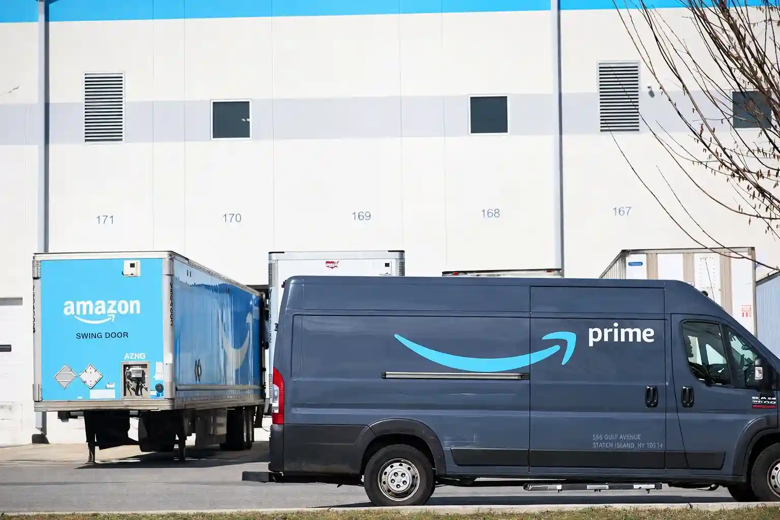 Amazon Prime delivery van parked outside a warehouse with Amazon-branded trailers in the background.