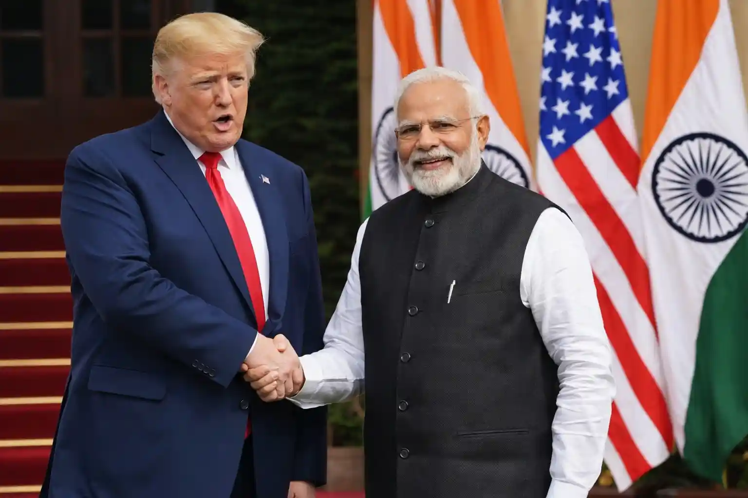 Former U.S. President Donald Trump shaking hands with Indian Prime Minister Narendra Modi with flags of India and the United States in the background.