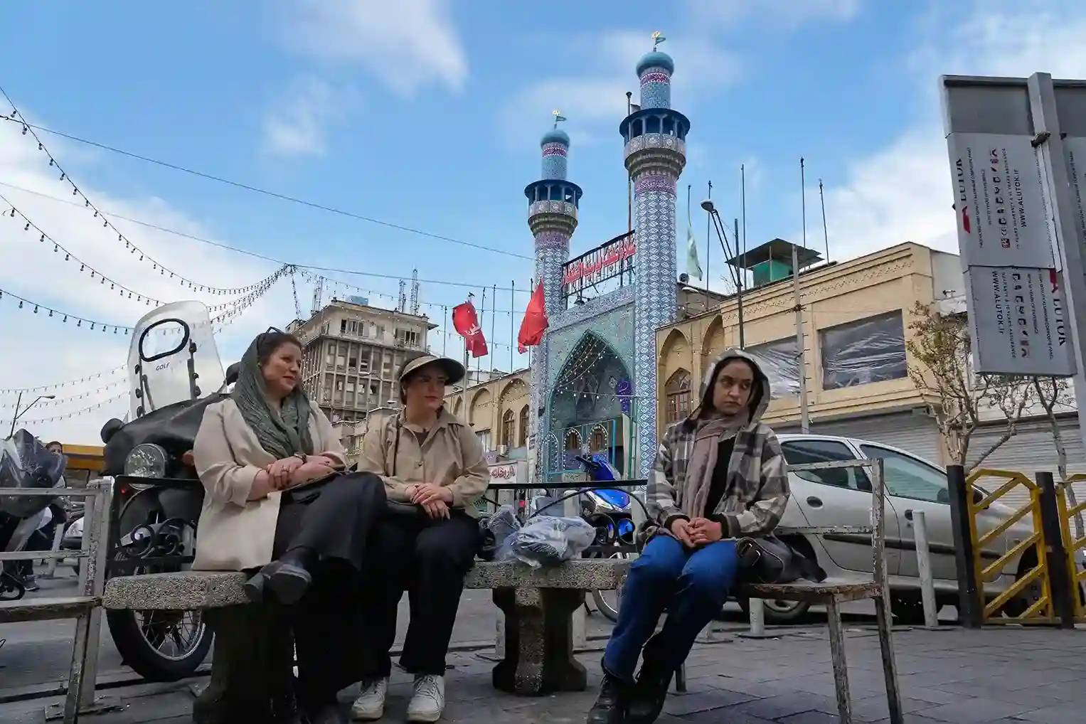 Women sitting on a street bench in an Iranian city with a colorful tiled mosque and minarets in the background