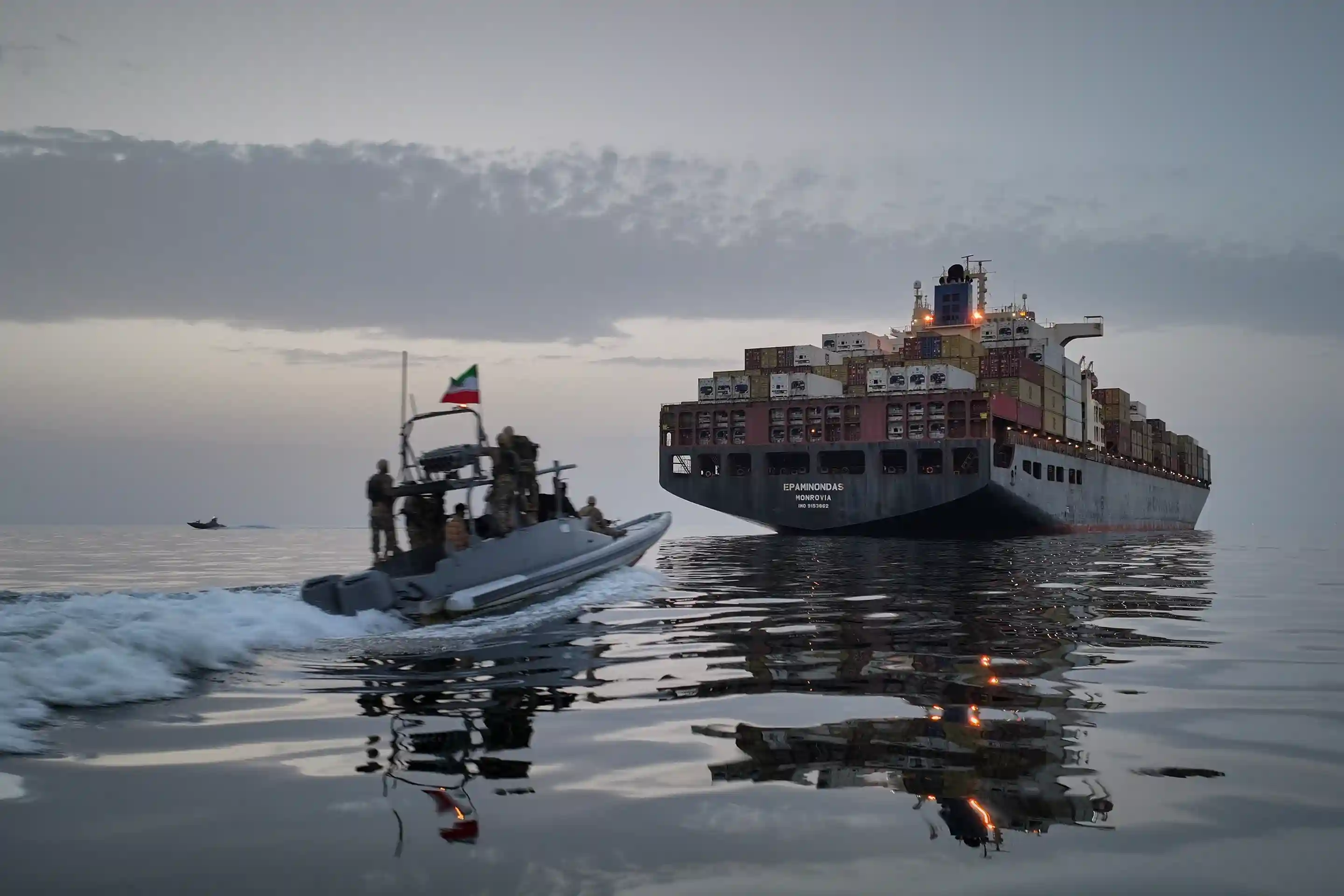 Armed patrol boat approaching a large container cargo ship in calm waters near the Strait of Hormuz