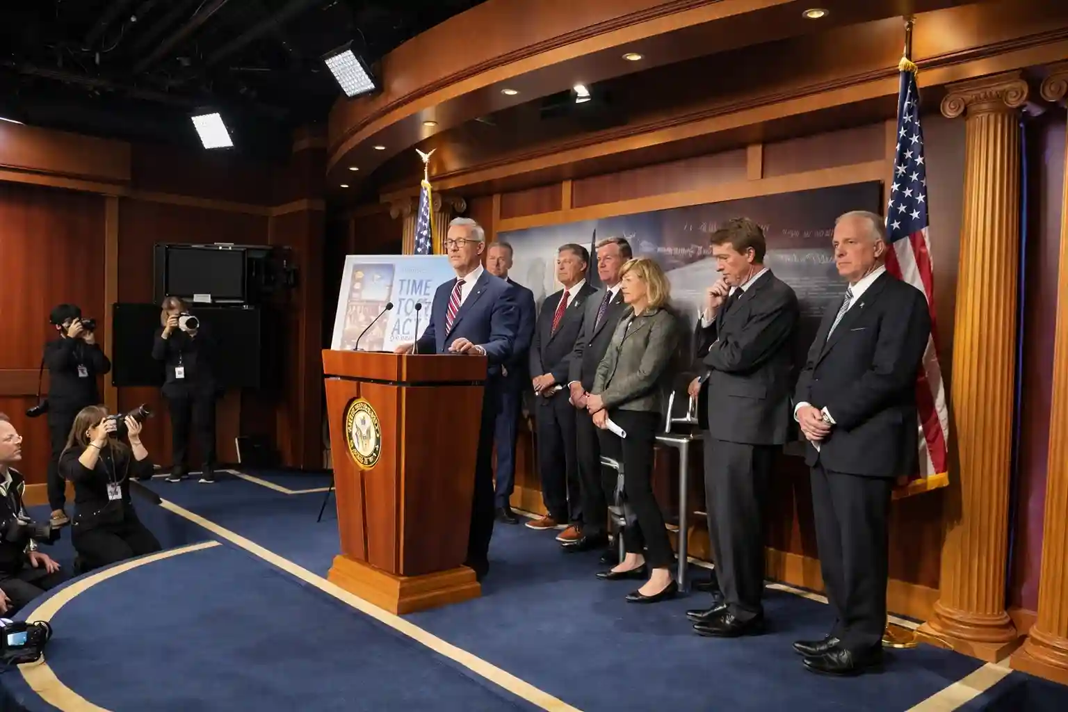 A press conference in a government setting with multiple officials standing behind a speaker, who is addressing the media.