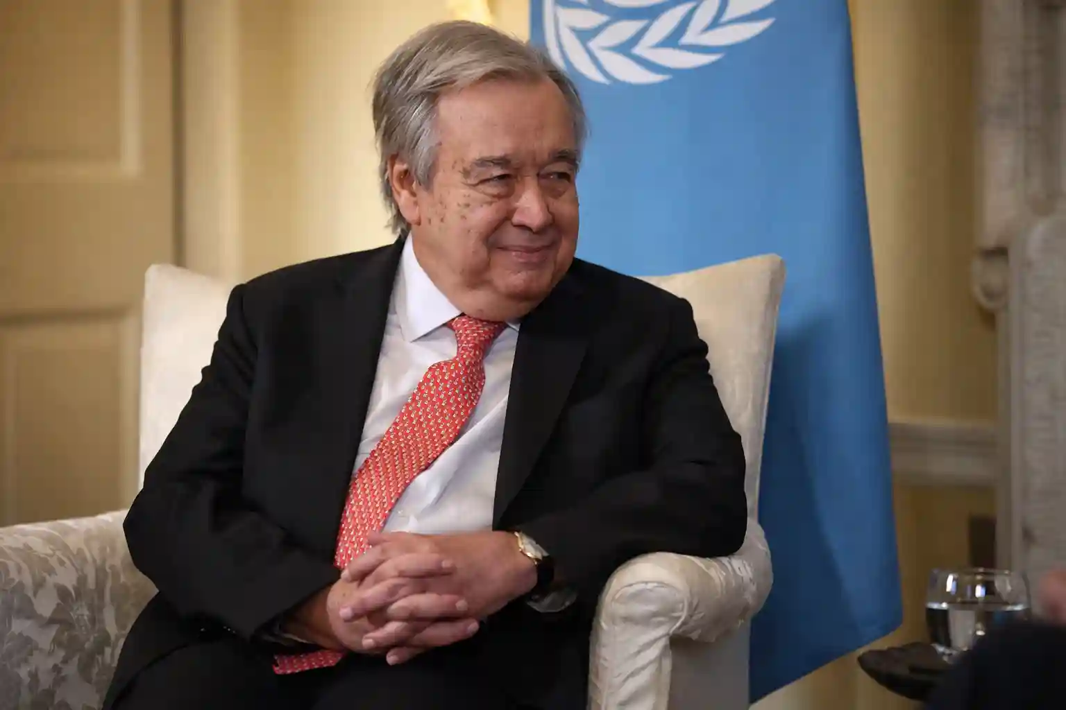 UN Secretary-General António Guterres smiling during an interview with a UN flag in the background.