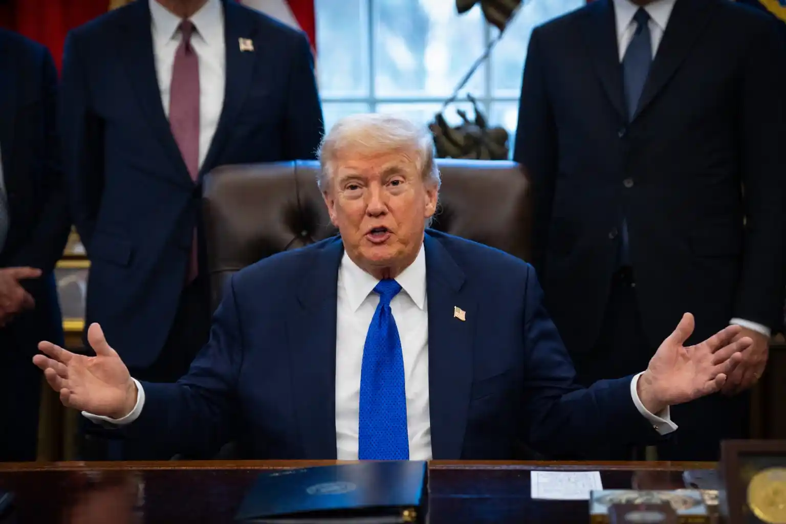 Donald Trump speaking in the Oval Office with hands raised, addressing an audience during a meeting.