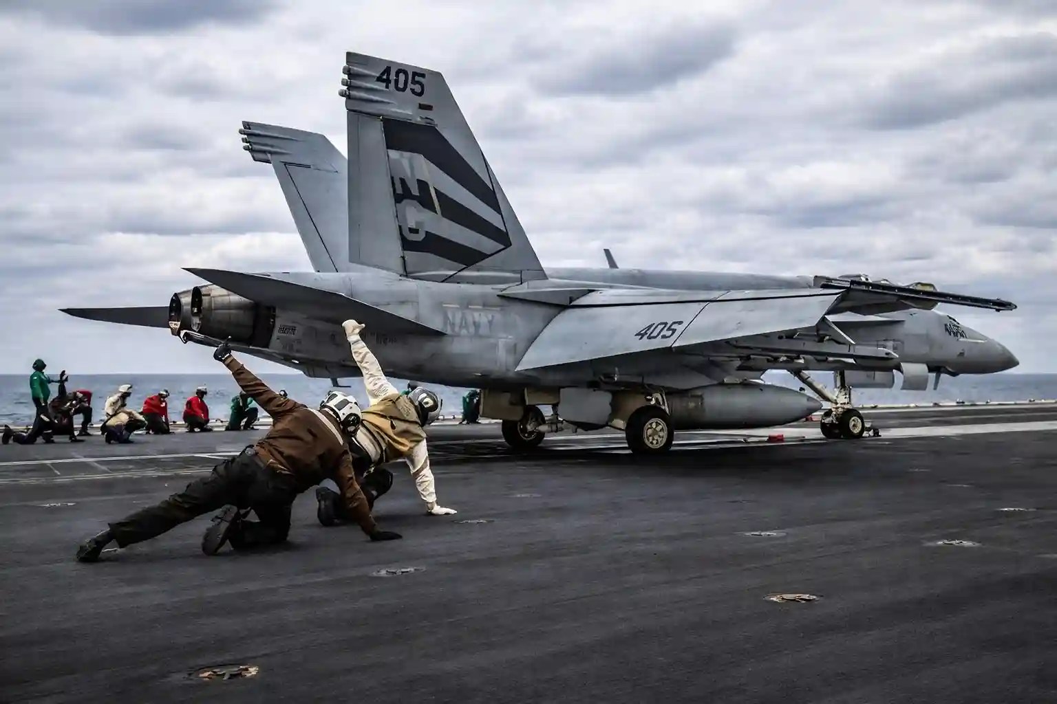 US Navy personnel performing a launch operation on an aircraft carrier, with an F/A-18 Hornet jet on the deck.