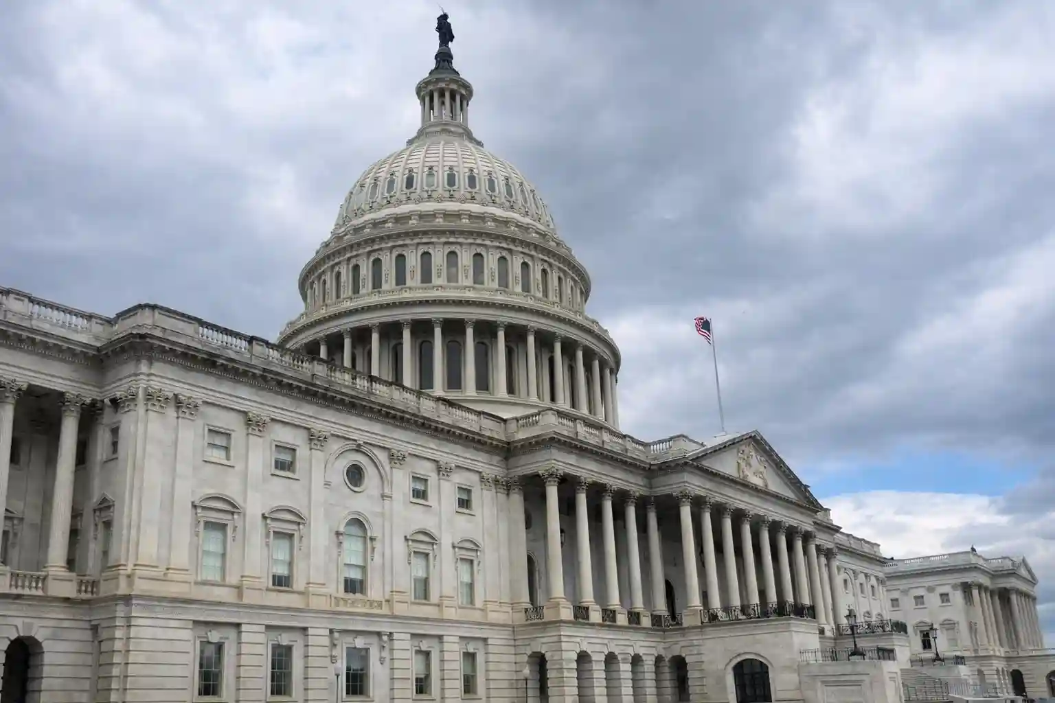 The U.S. Capitol building with the American flag flying on top, under a cloudy sky.