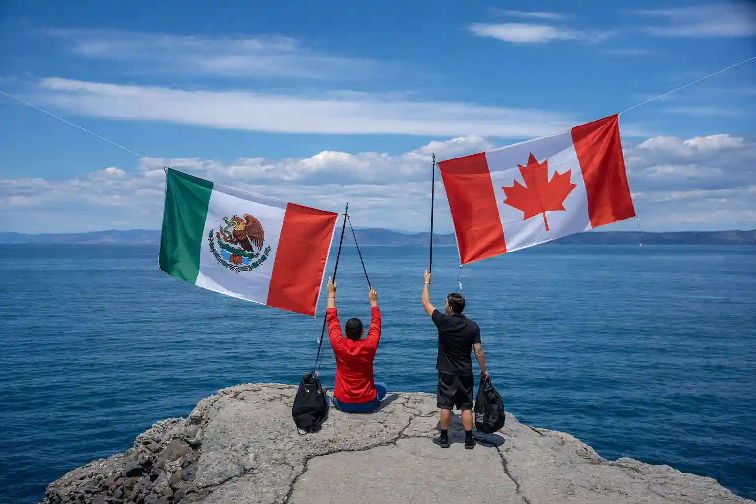 Two people holding the flags of Mexico and Canada on a rocky cliff overlooking the ocean.
